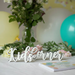 White decorative script sign reading 'Kids Corner' with black outline, displayed on a tabletop with flowers, balloons, and a notebook.